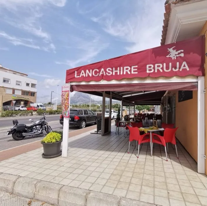 The Lancashire Bruja storefront — wide red awning with white witch-on-broomstick logo, terrace with yellow tables and red chairs, the Montgó mountain visible in the background, El Arenal, Jávea (Xàbia), Costa Blanca, Spain